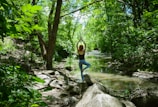 A person is practicing yoga on a large rock by a serene forest stream. Surrounded by lush, green foliage, the individual stands in a tree pose with arms raised, facing away. Sunlight filters through the tree canopy, casting dappled light on the forest floor.