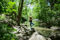 A person is practicing yoga on a large rock by a serene forest stream. Surrounded by lush, green foliage, the individual stands in a tree pose with arms raised, facing away. Sunlight filters through the tree canopy, casting dappled light on the forest floor.