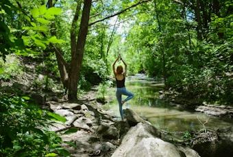 A person is practicing yoga on a large rock by a serene forest stream. Surrounded by lush, green foliage, the individual stands in a tree pose with arms raised, facing away. Sunlight filters through the tree canopy, casting dappled light on the forest floor.