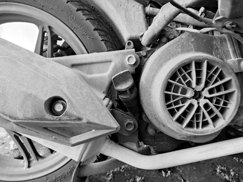 A close-up view of a motorcycle's mechanical parts, including the wheel with visible tire tread, a protective cover, and a circular fan-like component. The image is in black and white, highlighting the textures and grit on the metal surfaces.