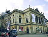 A historic building with classical architecture featuring arched windows and intricate carvings. There is scaffolding on one side, indicating construction or restoration work. Banners are displayed on the facade, and a statue is situated near the entrance. A truck and a cyclist are on the cobblestone street in front of the building.