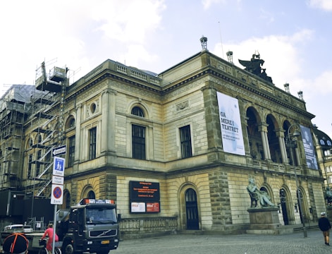 A historic building with classical architecture featuring arched windows and intricate carvings. There is scaffolding on one side, indicating construction or restoration work. Banners are displayed on the facade, and a statue is situated near the entrance. A truck and a cyclist are on the cobblestone street in front of the building.