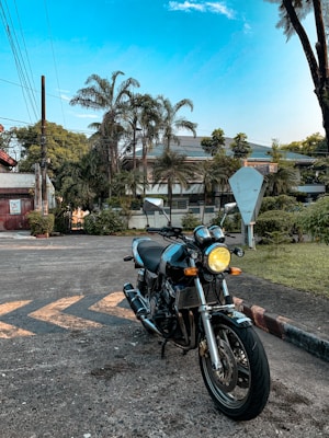 A classic motorcycle is parked on a paved road with palm trees and a house in the background. The motorcycle has dual headlights and a sleek design that stands out against the clear blue sky. The setting appears residential with a mix of greenery and urban elements.