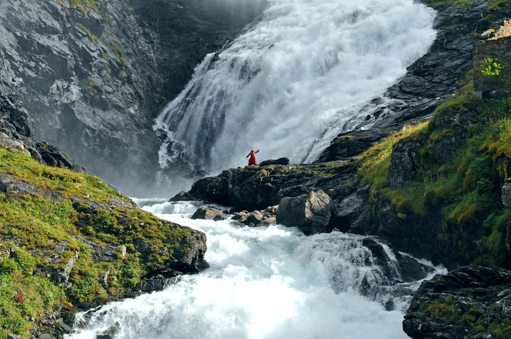 person in red jacket sitting on rock near waterfalls during daytime