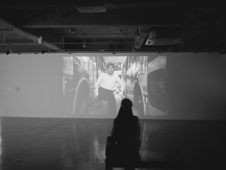 A person is seated on a bench in a dark room, viewing a large black and white projection on a wall. The projection depicts a man standing partially on a bus in an urban or street setting.