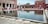A rectangular water basin is surrounded by a large courtyard with red sandstone architecture. Several people, including children, are walking and sitting around the basin. The arches in the background and the reflection in the water add to the architectural beauty of the scene.