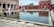 A rectangular water basin is surrounded by a large courtyard with red sandstone architecture. Several people, including children, are walking and sitting around the basin. The arches in the background and the reflection in the water add to the architectural beauty of the scene.