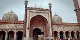 A large mosque with ornate architecture featuring multiple domes and minarets. The facade showcases detailed carvings and intricate patterns in red and white stone. Several people are seen in the foreground, including a man wearing traditional attire, suggesting a cultural or religious setting.