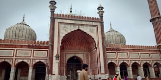A large mosque with ornate architecture featuring multiple domes and minarets. The facade showcases detailed carvings and intricate patterns in red and white stone. Several people are seen in the foreground, including a man wearing traditional attire, suggesting a cultural or religious setting.