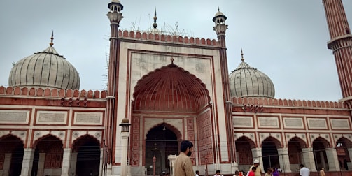 A large mosque with ornate architecture featuring multiple domes and minarets. The facade showcases detailed carvings and intricate patterns in red and white stone. Several people are seen in the foreground, including a man wearing traditional attire, suggesting a cultural or religious setting.
