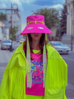 A person wearing a vibrant, oversized neon green jacket with fringes and matching bright pink hat that covers part of the face. The hat has eye graphics and long straps with text. The shirt underneath has a colorful, playful design. The background features an urban street scene with a blurred cityscape, including buildings and parked cars.