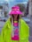 A person wearing a vibrant, oversized neon green jacket with fringes and matching bright pink hat that covers part of the face. The hat has eye graphics and long straps with text. The shirt underneath has a colorful, playful design. The background features an urban street scene with a blurred cityscape, including buildings and parked cars.