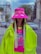 A person wearing a vibrant, oversized neon green jacket with fringes and matching bright pink hat that covers part of the face. The hat has eye graphics and long straps with text. The shirt underneath has a colorful, playful design. The background features an urban street scene with a blurred cityscape, including buildings and parked cars.