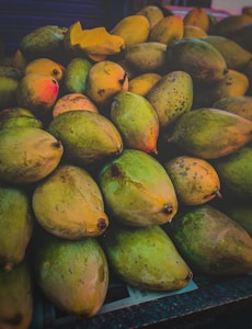 A pile of ripe and unripe mangoes with varying shades of green and yellow, some displaying spots. The lighting is moody with shadows cast over the fruit, and a single cut mango slice is visible amidst the whole fruits.
