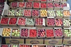 A variety of fruits are neatly packed in cardboard boxes, each containing a different kind such as apples, plums, pears, and tomatoes. The boxes are arranged in rows, showcasing their vibrant colors and freshness.