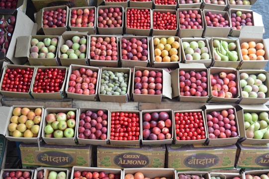A variety of fruits are neatly packed in cardboard boxes, each containing a different kind such as apples, plums, pears, and tomatoes. The boxes are arranged in rows, showcasing their vibrant colors and freshness.