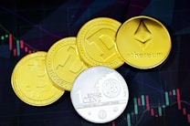 A collection of four coin-like representations of cryptocurrencies including Ethereum, Bitcoin, and Dogecoin. Each coin displays symbols or text related to its respective cryptocurrency. The background features a financial chart with candlestick patterns, suggesting a trading or investment context.