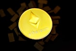 A gold coin with the Ethereum logo embossed on it is placed against a dark background with scattered black rectangles around it. The word 'ethereum' is inscribed beneath the logo.