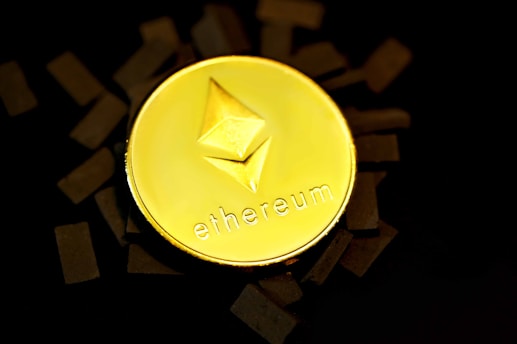 A gold coin with the Ethereum logo embossed on it is placed against a dark background with scattered black rectangles around it. The word 'ethereum' is inscribed beneath the logo.