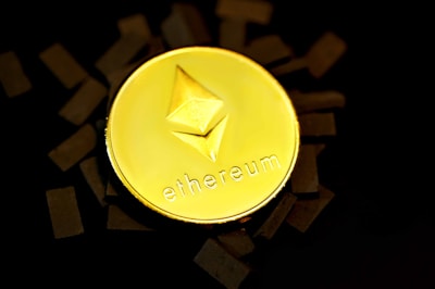 A gold coin with the Ethereum logo embossed on it is placed against a dark background with scattered black rectangles around it. The word 'ethereum' is inscribed beneath the logo.