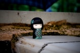 A small, stylized doll with a black and green design stands on a rough, uneven surface. The doll has a serene expression with closed eyes and red lips, complemented by an intricate floral pattern on its clothing. The background is a blurred mix of natural tones and textures, possibly including foliage and stone.