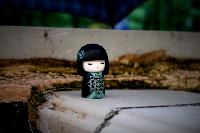 A small, stylized doll with a black and green design stands on a rough, uneven surface. The doll has a serene expression with closed eyes and red lips, complemented by an intricate floral pattern on its clothing. The background is a blurred mix of natural tones and textures, possibly including foliage and stone.