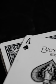 A close-up of playing cards featuring the ace of spades with intricate black and white designs. The card displays the word 'Bicycle' and includes detailed artwork on a dark background.