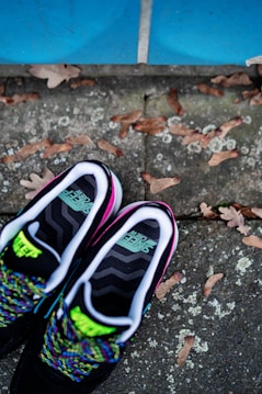 A pair of black sneakers with colorful laces is placed on a textured concrete surface with dry leaves scattered around. The sneakers have a vibrant design with neon green accents and text visible on the insoles. Above, there is a blue tiled section, creating a contrasting background.