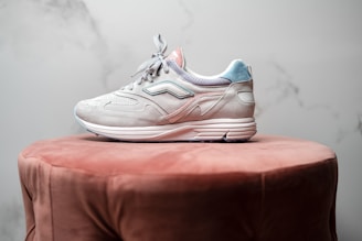 A single sneaker with a light gray and pastel blue color scheme is placed on a plush, brown circular ottoman. The shoe features intricate stitching and a sleek design, with a textured sole that suggests a focus on style and comfort. The background is a soft marble texture in muted tones.