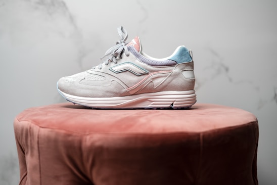 A single sneaker with a light gray and pastel blue color scheme is placed on a plush, brown circular ottoman. The shoe features intricate stitching and a sleek design, with a textured sole that suggests a focus on style and comfort. The background is a soft marble texture in muted tones.