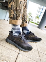A person wearing camo-patterned pants and stylish dark sneakers with a logo on a paved outdoor area. The lower legs are visible, and the background features trees and a multi-story building.