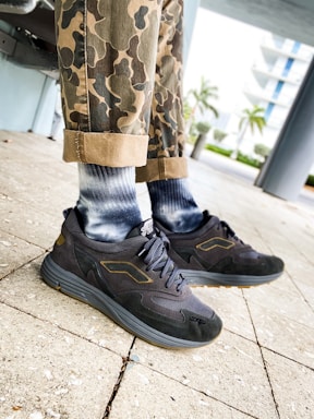 A person wearing camo-patterned pants and stylish dark sneakers with a logo on a paved outdoor area. The lower legs are visible, and the background features trees and a multi-story building.