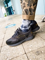 A close-up of a person's lower leg wearing camouflage-patterned pants rolled up at the cuffs, tie-dye socks in shades of white and blue, and black athletic shoes. The background shows a tiled floor with some blue social distancing markers and a modern glass storefront.