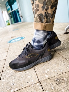 A close-up of a person's lower leg wearing camouflage-patterned pants rolled up at the cuffs, tie-dye socks in shades of white and blue, and black athletic shoes. The background shows a tiled floor with some blue social distancing markers and a modern glass storefront.