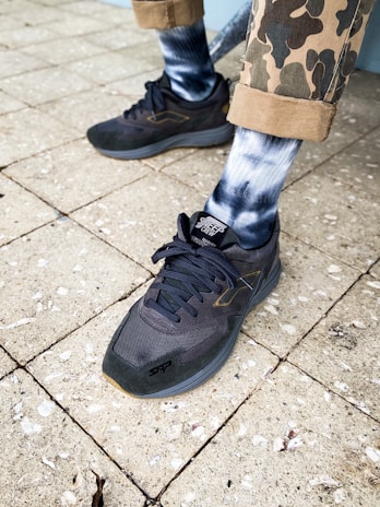 A pair of feet wearing dark sneakers, patterned socks, and camo-style rolled pants stand on a textured, tiled pavement.