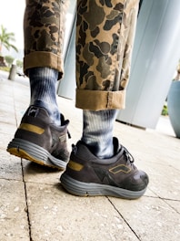 A pair of legs wearing camouflage pants with the cuffs rolled up, revealing blue and white gradient socks, are shown walking on a tiled pavement. The person is wearing black and gray sneakers with yellow accents. In the background, there is a large cylindrical structure and some potted plants.
