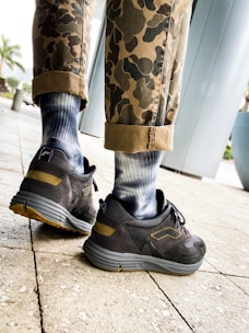 A pair of legs wearing camouflage pants with the cuffs rolled up, revealing blue and white gradient socks, are shown walking on a tiled pavement. The person is wearing black and gray sneakers with yellow accents. In the background, there is a large cylindrical structure and some potted plants.