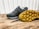 A pair of dark-colored athletic shoes placed on a wooden surface, with one shoe laying on its side to display the sole. The shoes have a sleek design with a mix of suede and mesh materials, featuring a rubber sole with unique tread patterns for grip.
