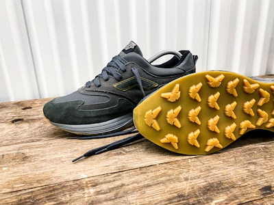 A pair of dark-colored athletic shoes placed on a wooden surface, with one shoe laying on its side to display the sole. The shoes have a sleek design with a mix of suede and mesh materials, featuring a rubber sole with unique tread patterns for grip.