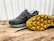 A pair of dark-colored athletic shoes placed on a wooden surface, with one shoe laying on its side to display the sole. The shoes have a sleek design with a mix of suede and mesh materials, featuring a rubber sole with unique tread patterns for grip.