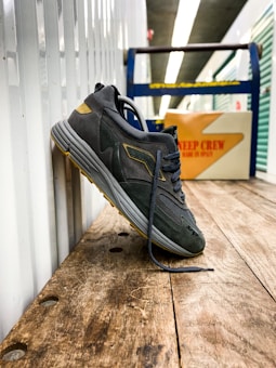 A single dark-colored sneaker with gray and yellow accents is leaning against a corrugated metal wall on a wooden surface. The shoe is upright, with its laces undone and loosely hanging. In the background, a cardboard box is visible, displaying the text 'DEEP CREW MADE IN SPAIN' in red. The setting appears to be an indoor storage area or workshop, with blurred shelving in the background.