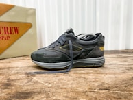 A stylish athletic shoe positioned on a wooden surface, with a mix of suede and mesh materials in dark tones. The shoe features intricate detailing with a distinctive logo on the side. In the background, there is a vintage-style box with bold text featuring the word 'SPAIN'.