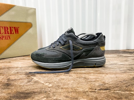 A stylish athletic shoe positioned on a wooden surface, with a mix of suede and mesh materials in dark tones. The shoe features intricate detailing with a distinctive logo on the side. In the background, there is a vintage-style box with bold text featuring the word 'SPAIN'.