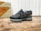 A stylish athletic shoe positioned on a wooden surface, with a mix of suede and mesh materials in dark tones. The shoe features intricate detailing with a distinctive logo on the side. In the background, there is a vintage-style box with bold text featuring the word 'SPAIN'.