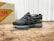 A stylish athletic shoe positioned on a wooden surface, with a mix of suede and mesh materials in dark tones. The shoe features intricate detailing with a distinctive logo on the side. In the background, there is a vintage-style box with bold text featuring the word 'SPAIN'.