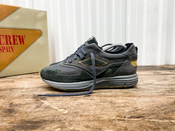 A stylish athletic shoe positioned on a wooden surface, with a mix of suede and mesh materials in dark tones. The shoe features intricate detailing with a distinctive logo on the side. In the background, there is a vintage-style box with bold text featuring the word 'SPAIN'.