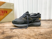 A stylish athletic shoe positioned on a wooden surface, with a mix of suede and mesh materials in dark tones. The shoe features intricate detailing with a distinctive logo on the side. In the background, there is a vintage-style box with bold text featuring the word 'SPAIN'.