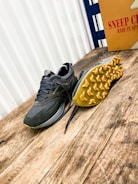 A pair of athletic shoes with a rugged sole is placed on a wooden surface. The shoe on the left is upright, while the one on the right is flipped to display the yellow tread pattern. A cardboard box with the text 'Sneep Crew Made in Spain' is visible in the background.