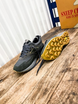 A pair of athletic shoes with a rugged sole is placed on a wooden surface. The shoe on the left is upright, while the one on the right is flipped to display the yellow tread pattern. A cardboard box with the text 'Sneep Crew Made in Spain' is visible in the background.