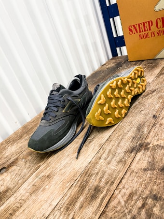 A pair of athletic shoes with a rugged sole is placed on a wooden surface. The shoe on the left is upright, while the one on the right is flipped to display the yellow tread pattern. A cardboard box with the text 'Sneep Crew Made in Spain' is visible in the background.